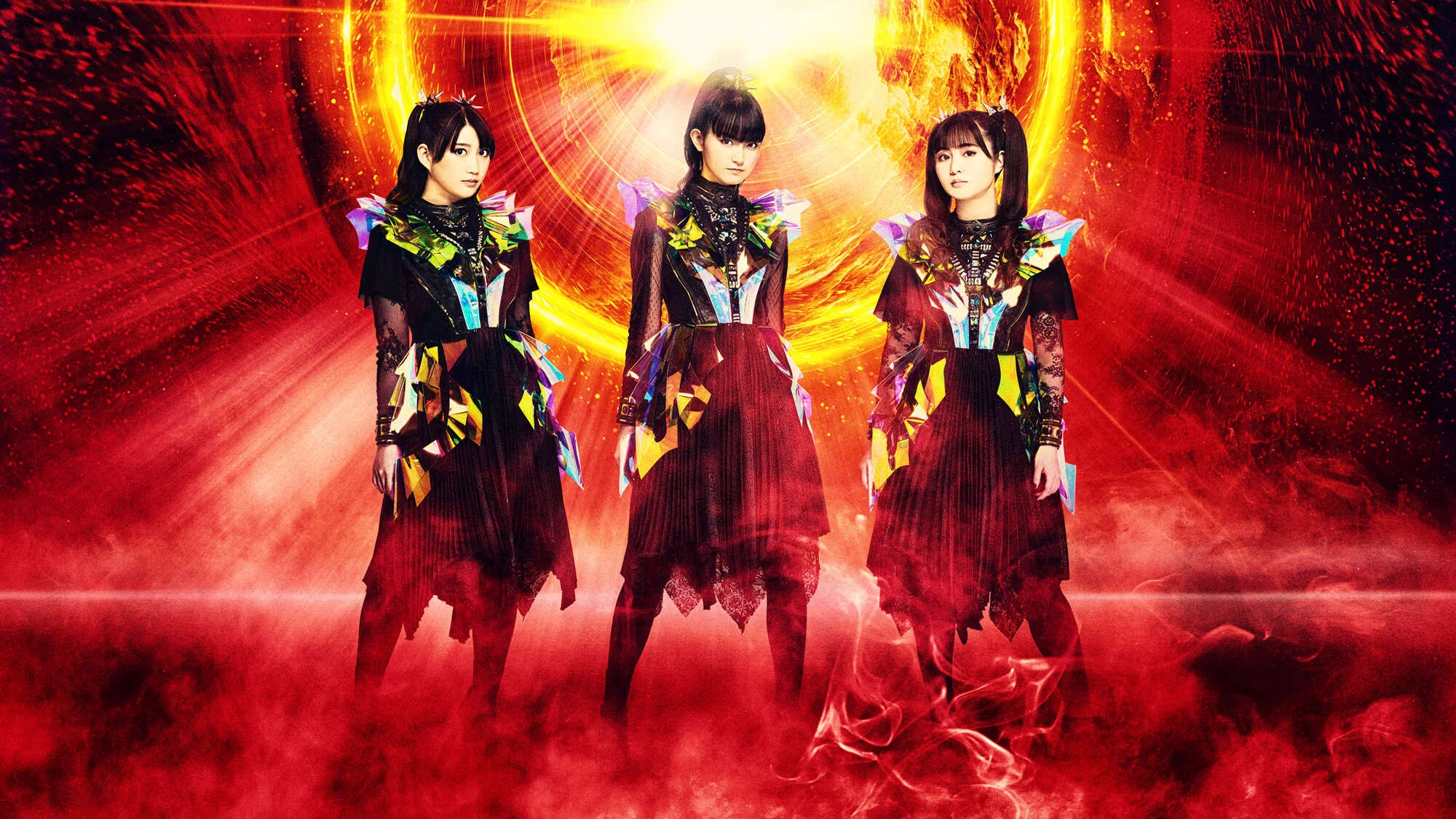 BABYMETAL frente a un anillo dorado gigante y resplandeciente, sobre un fondo con rayos de luces rojas