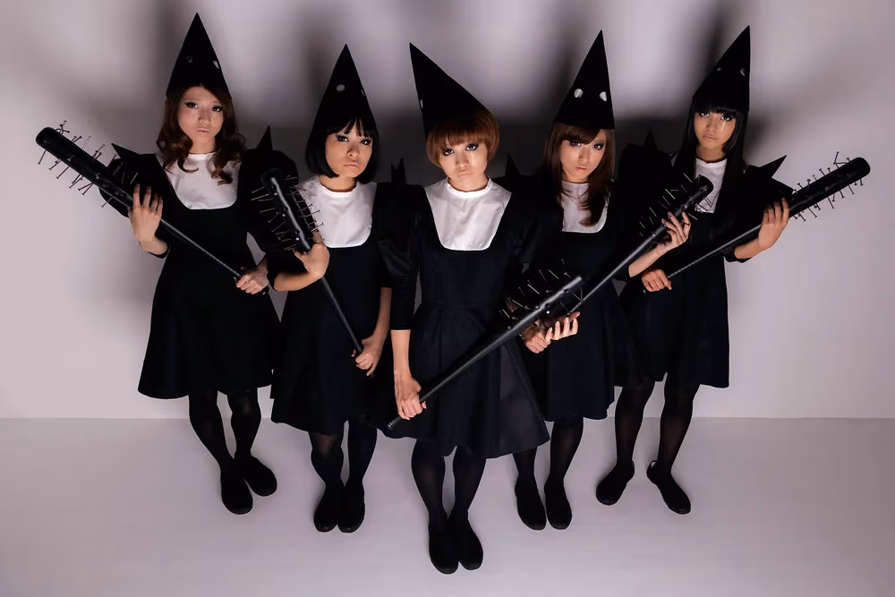 Las 5 chicas de BiS, formadas en línea en un cuarto blanco, mirando a la cámara y llevando vestidos de cuello blanco, sombreros cónicos y bates de púas, todos de color negro